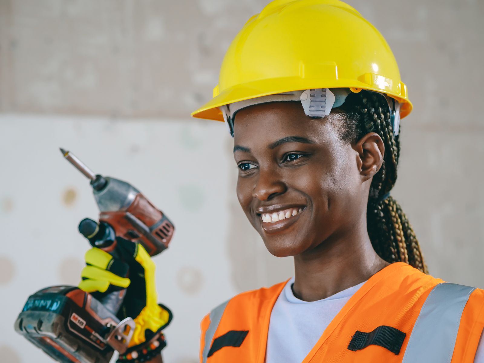 A young worker in a yellow hard hat and orange vest smiling while holding a drill